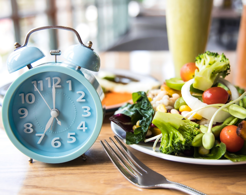 Assiette de légumes colorée sur une table en bois à côté d'un réveil bleu indiquant 7 heures du matin